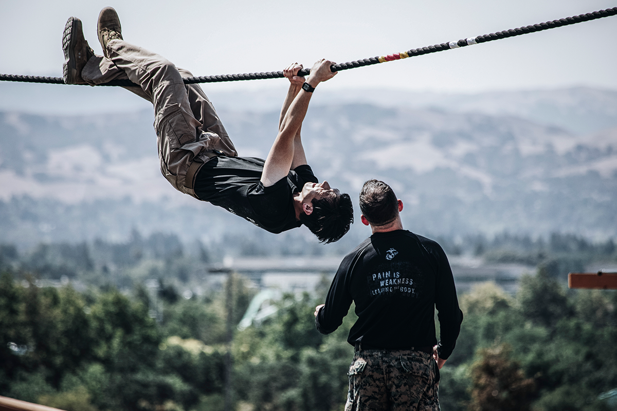 Officer candidate performs inverted rope climb exercise while instructor observes during physical fitness training.