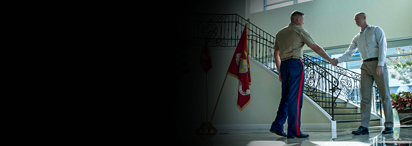 Officer in dress uniform shakes hands with prospective candidate in modern building with battle colors displayed.