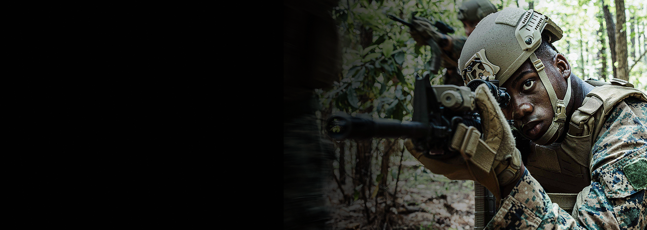 A Marine win camouflage looks down the scope of his rifle as he kneels in the woods. 
