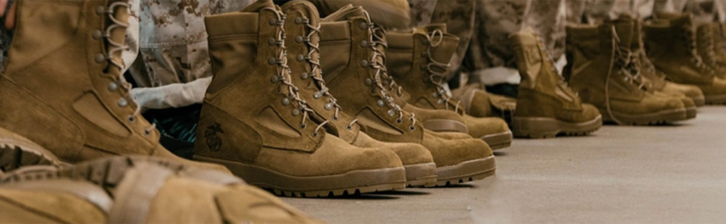 Multiple pairs of tan Marine combat boots with laces, one featuring the Marine Corps emblem, lined up on a dark surface.