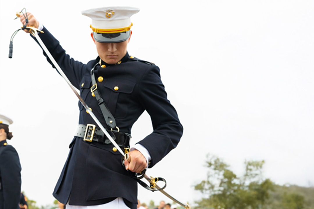 A Marine in dress uniform sheathing a mameluke sword, looking down, with a blurry outdoor background.