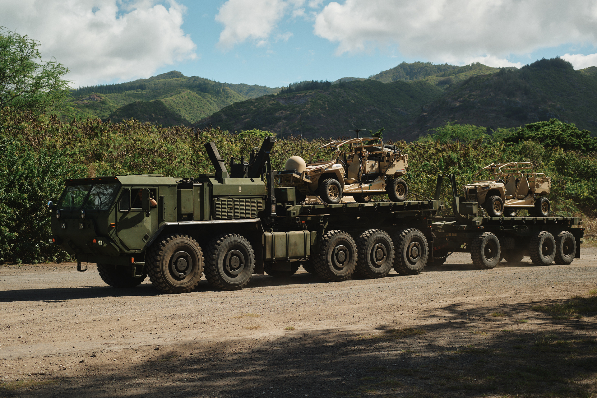 Heavy equipment transport truck carries multiple tactical vehicles on flatbed trailer in Hawaii mountain terrain.