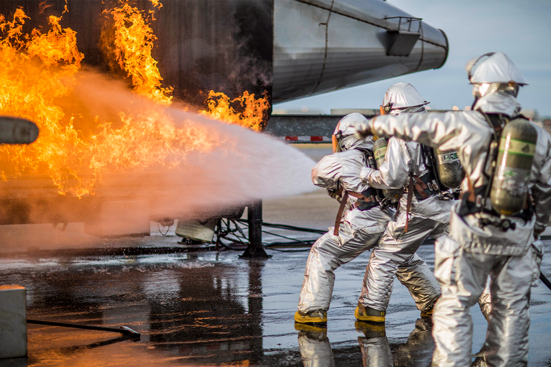 Marines in silver firefighting suits and oxygen tanks put of a training fire with large houses on the tarmac of an airfield. 