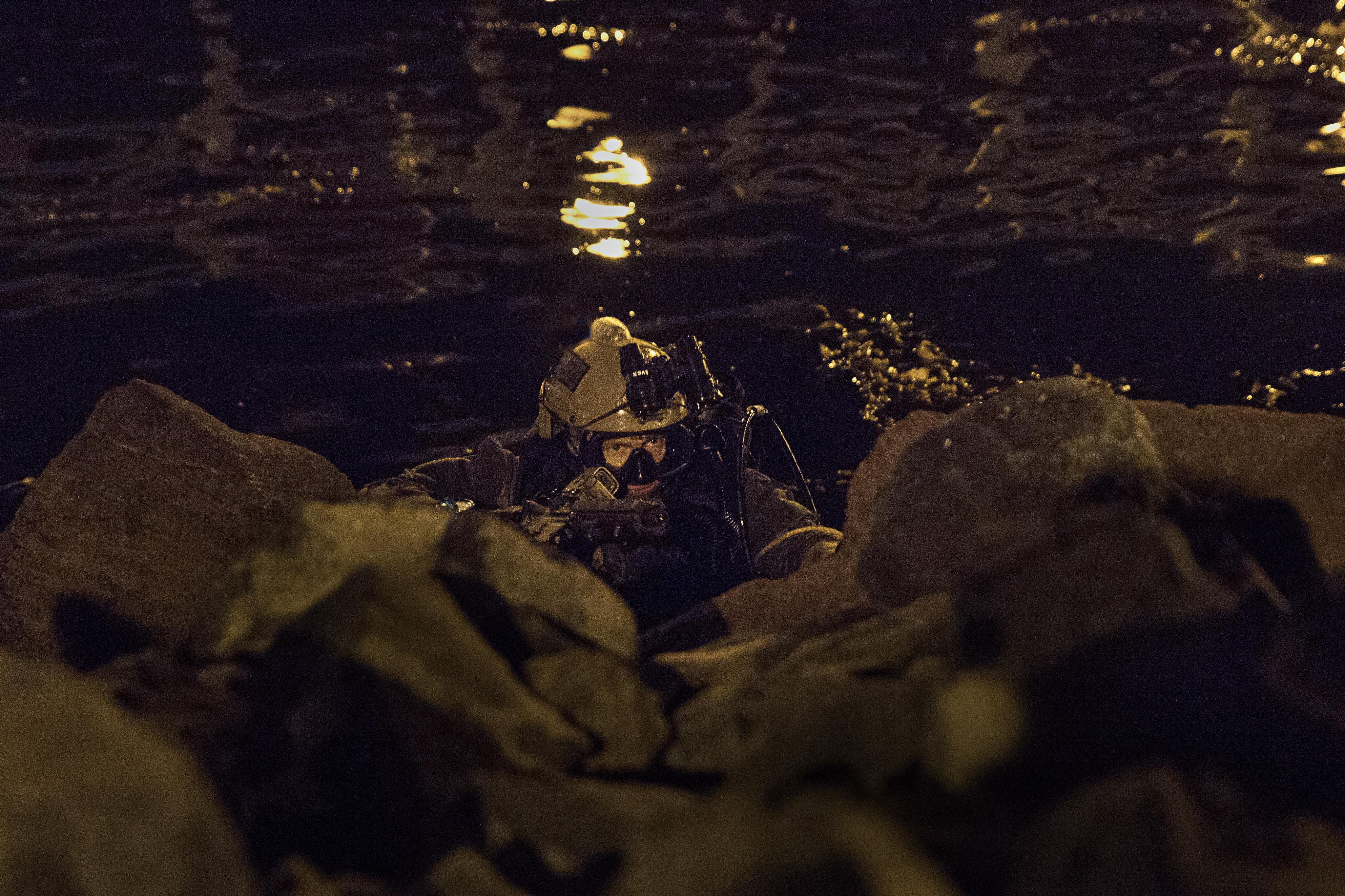 A special operations Marine in goggles and scuba gear rises out of the water at night. 
