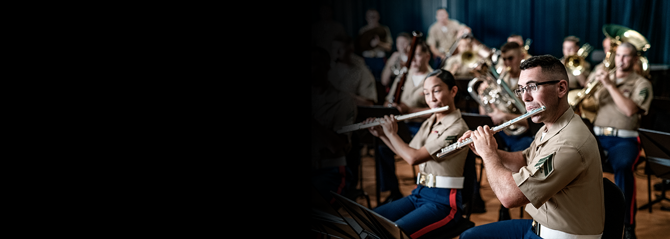 Marine musicians in service uniforms play flutes and brass instruments during indoor rehearsal.