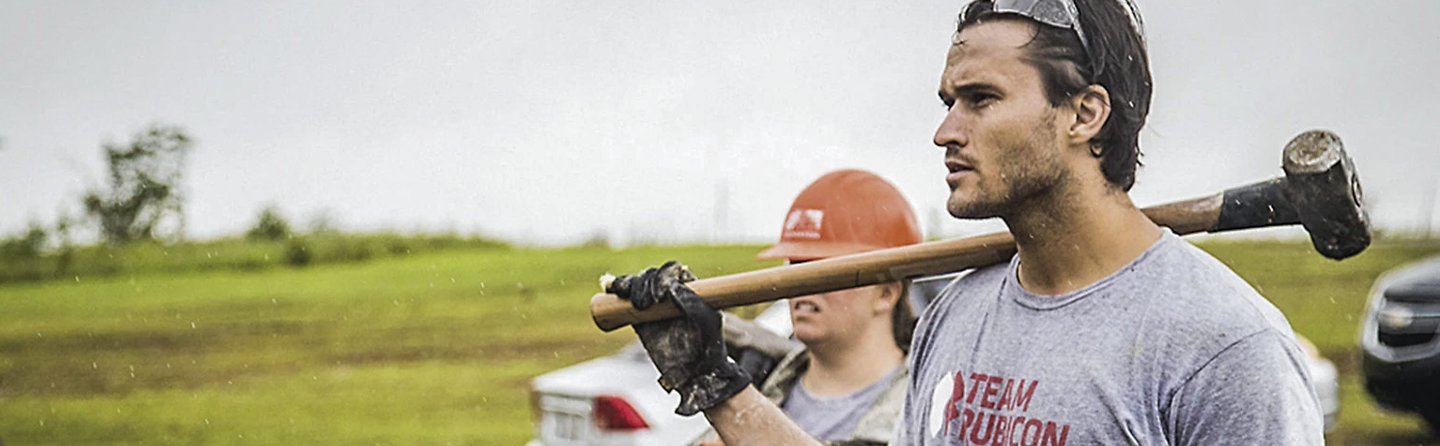 Team Rubicon's Jake Wood carries sledgehammer over shoulder during disaster relief operation in field setting.